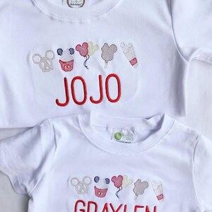 May include: Two white t-shirts with embroidered designs and red text. The top shirt says "JOJO" and the bottom shirt says "GRAYLEN". Both shirts feature a row of embroidered images including a pretzel, cupcake, balloons, ice cream, and a popsicle.