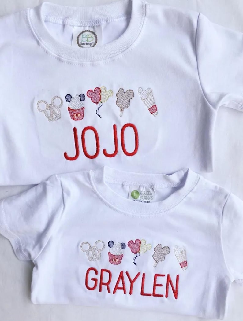 May include: Two white t-shirts with embroidered designs and red text. The top shirt says "JOJO" and the bottom shirt says "GRAYLEN". Both shirts feature a row of embroidered images including a pretzel, cupcake, balloons, ice cream, and a popsicle.