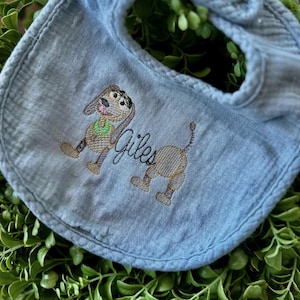 May include: A light blue baby bib with an embroidered cartoon dog design. The dog is brown and tan with a green collar. The name "Giles" is embroidered in cursive below the dog.