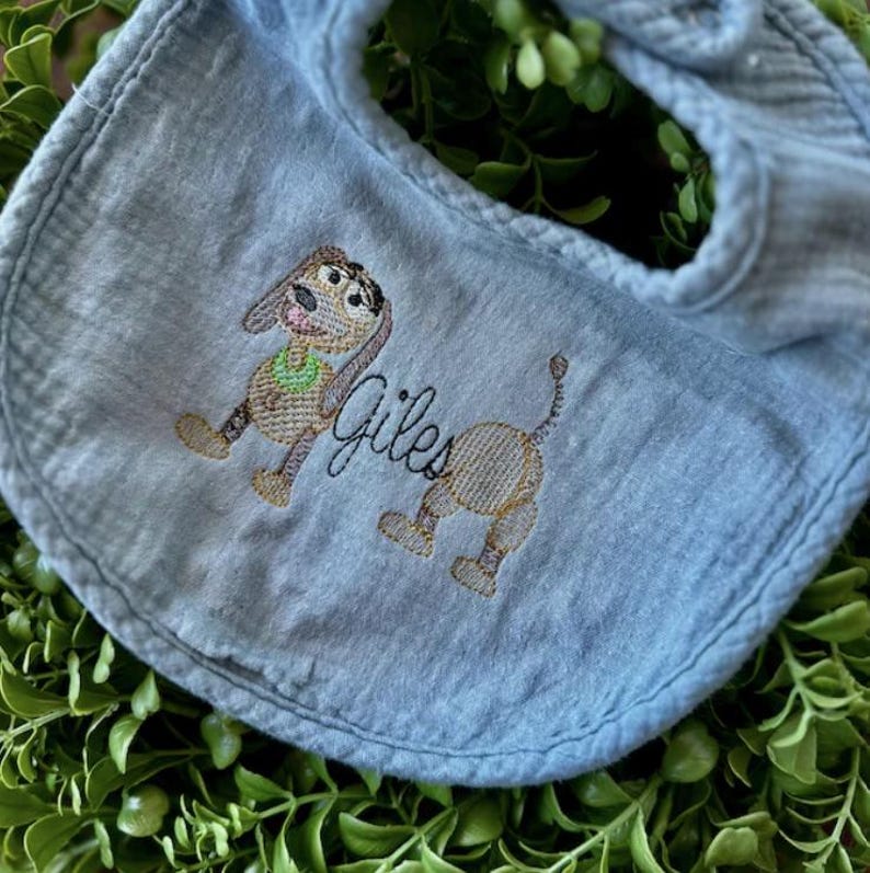 May include: A light blue baby bib with an embroidered cartoon dog design. The dog is brown and tan with a green collar. The name "Giles" is embroidered in cursive below the dog.