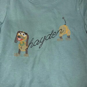 May include: A light teal-colored shirt with an embroidered dog design and the name "Hayden" in cursive. The dog is yellow, brown, and black, with a smiling face. The design is on the front of the shirt.