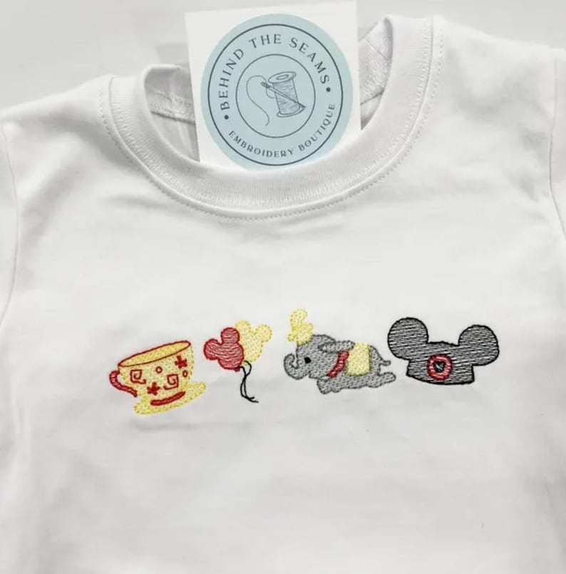 May include: White baby onesie featuring embroidered images of a teacup, balloons, an elephant, and a mouse head. A tag with the text "BEHIND THE SEAMS EMBROIDERY BOUTIQUE" is attached to the neckline.