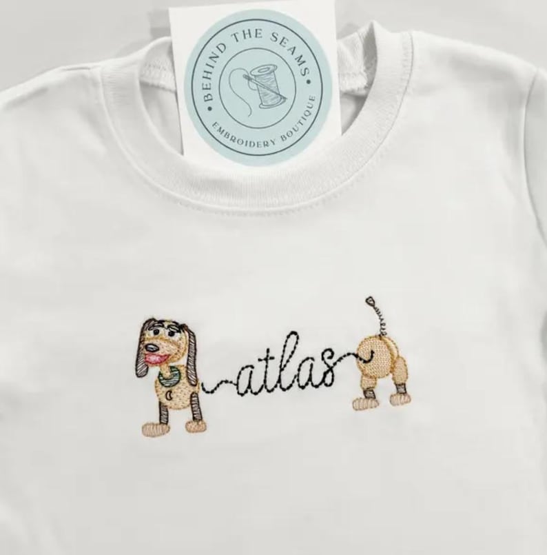 May include: White t-shirt with an embroidered dog design and the name "atlas". The dog is brown, tan, and black. A tag at the top reads "BEHIND THE SEAMS EMBROIDERY BOUTIQUE" in a circular design.