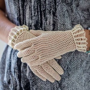 Puede incluir: Guantes color crema, de tejido abierto con un delicado borde festoneado. Los guantes est&aacute;n hechos de un material fino, similar a una red, y el dise&ntilde;o permite que la piel sea visible. Los guantes son de estilo vintage y el aspecto general es elegante y refinado.