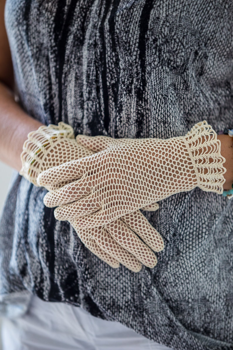 Puede incluir: Guantes color crema, de tejido abierto con un delicado borde festoneado. Los guantes est&aacute;n hechos de un material fino, similar a una red, y el dise&ntilde;o permite que la piel sea visible. Los guantes son de estilo vintage y el aspecto general es elegante y refinado.
