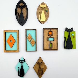 May include: A collection of eight wooden wall art pieces featuring cat silhouettes, geometric shapes, and starburst accents. The items include various shapes like ovals, rectangles, and diamonds, finished in colors including black, gold, orange, and teal.