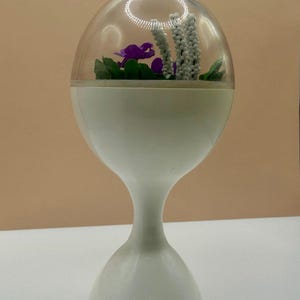 May include: A white pedestal stand with a clear, dome-shaped top containing a miniature arrangement of purple flowers and white beaded stalks. The base is hourglass-shaped, and the clear dome features a circular light reflection at the top.