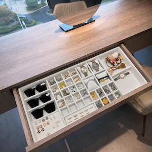 Op de afbeelding: Een houten lade van een kaptafel met een witte fluwelen sieradenorganizer. De lade bevat drie paar zwarte zonnebrillen, diverse gouden en zilveren oorbellen, meerdere ringen, twee polshorloges op kussentjes, een armband en haaraccessoires.