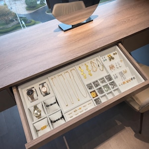 May include: A wooden vanity drawer featuring a white velvet jewelry organizer insert. The tray contains four wristwatches on cushions, two beaded bracelets, several gold-tone necklaces, various pairs of earrings, and multiple rings.