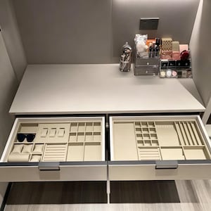 May include: A white vanity table with two open drawers featuring beige velvet jewelry organizers. The left drawer contains watch pillows and sunglasses, while the right drawer has various compartments. A clear acrylic makeup organizer sits on top.