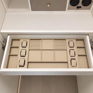 May include: A white drawer organizer insert featuring a beige velvet interior with multiple compartments. It includes six watch pillows, two ring storage sections with slots, and several open rectangular trays for organizing jewelry and accessories.