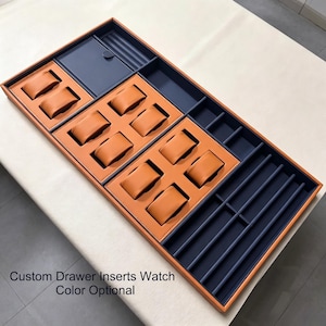 May include: A rectangular drawer organizer tray featuring a tan leather exterior and a dark blue interior. It includes ten tan watch pillows, a lidded compartment, and multiple long, narrow slots for storage. Text reads: Custom Drawer Inserts Watch Color Optional.