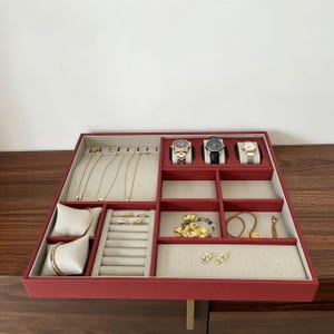 May include: A rectangular jewelry organizer tray with a deep red exterior and beige interior lining. It features dedicated compartments for watches, necklaces, rings, and earrings, including two beige velvet pillows for bracelets or watches.