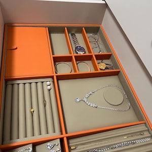 May include: An open drawer with an orange and beige jewelry organizer. The organizer holds a watch, glasses, rings, earrings, necklaces, and bracelets. The drawer is inside a white cabinet with a gold knob.