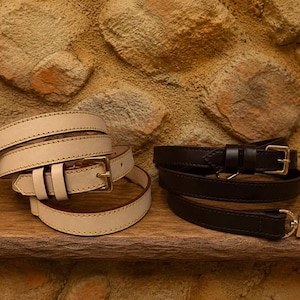 May include: Two leather belts, one cream-colored and one dark brown, are displayed on a wooden surface. The cream belt has a gold-colored buckle, and the dark brown belt has a silver-colored buckle. Both belts are rolled up.