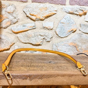 Pu&ograve; includere: Una tracolla per borsa color giallo senape con bordi lisci e arrotondati, dotata di chiusure a moschettone in metallo dorato su entrambe le estremit&agrave;. Il cinturino presenta cuciture visibili nei punti di attacco per un fissaggio sicuro.