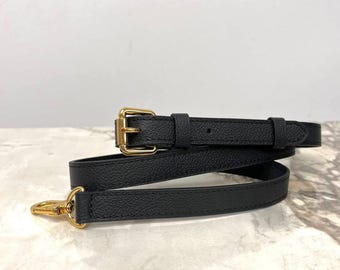 May include: A black textured leather bag strap featuring gold-toned metal hardware, including an adjustable buckle with a pin and a swivel lobster clasp hook at one end. The strap has a consistent width and a pebbled finish throughout its length.