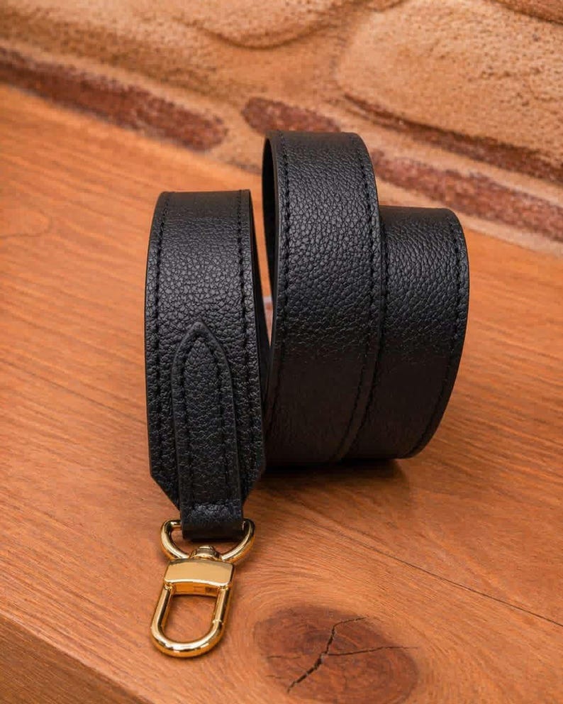 May include: A black leather strap with a gold-colored clasp. The strap is made of textured leather and has visible stitching along the edges. The clasp is a shiny gold color.