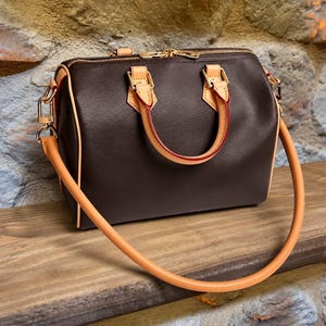 Pu&ograve; includere: Una borsa a mano marrone scuro a forma di barile con manici e finiture in pelle color cuoio chiaro, dettagli in metallo dorato, una chiusura superiore con zip e una tracolla lunga rimovibile con inserti in pelle color cuoio chiaro.