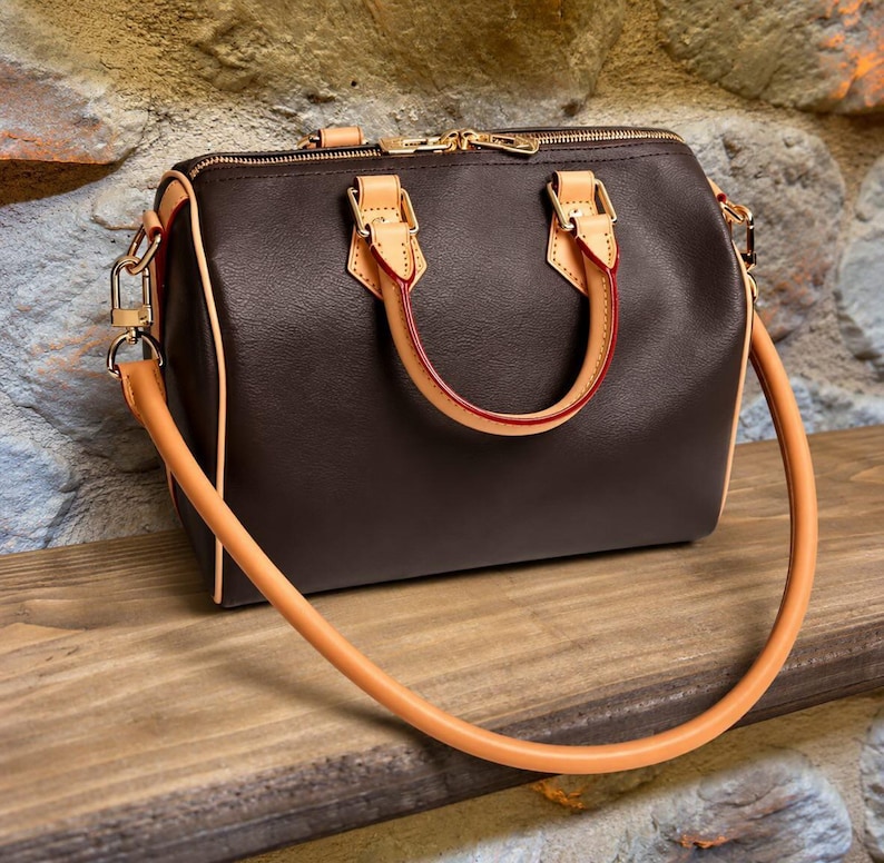 Pu&ograve; includere: Una borsa a mano marrone scuro a forma di barile con manici e finiture in pelle color cuoio chiaro, dettagli in metallo dorato, una chiusura superiore con zip e una tracolla lunga rimovibile con inserti in pelle color cuoio chiaro.
