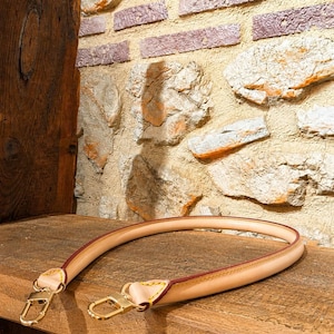 Pu&ograve; includere: Una tracolla per borsa in pelle color beige chiaro con bordi arrotondati e rifiniture dipinte in marrone scuro. Il cinturino presenta cuciture giallo brillante nei punti di attacco e due moschettoni in metallo dorato lucido.