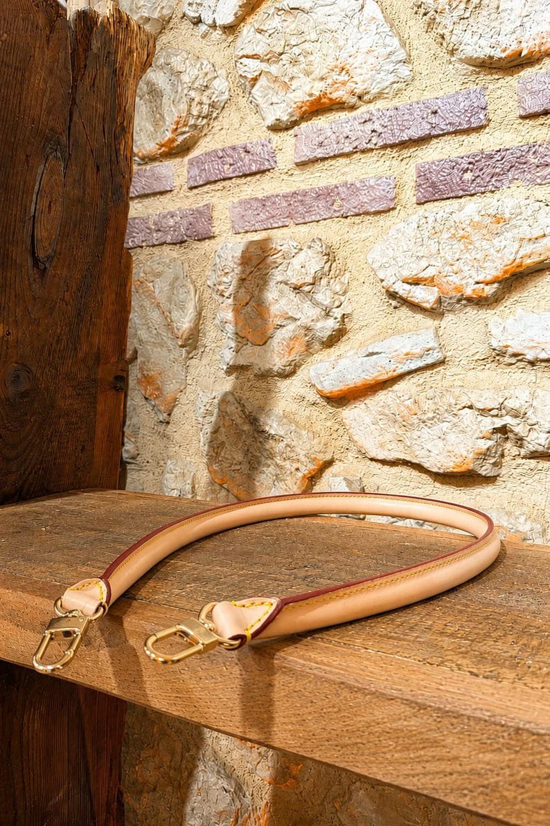 Pu&ograve; includere: Una tracolla per borsa in pelle color beige chiaro con bordi arrotondati e rifiniture dipinte in marrone scuro. Il cinturino presenta cuciture giallo brillante nei punti di attacco e due moschettoni in metallo dorato lucido.