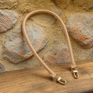 Può includere: Un manico per borsa in pelle beige con chiusure in metallo color oro. Il manico è curvo e ha una consistenza liscia. Le chiusure sono attaccate alle estremità del manico e sono progettate per essere attaccate a una borsa.