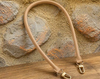 May include: A beige, rounded handbag strap featuring a smooth, tubular design. The strap is equipped with polished gold-tone metal lobster claw clasps at both ends, allowing for easy attachment to a bag. The strap has a consistent, clean finish.