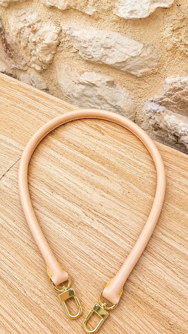 Pu&ograve; includere: Un manico per borsa in pelle color beige chiaro di forma arrotondata, dotato di chiusure a moschettone in metallo dorato su entrambe le estremit&agrave;. Il manico presenta cuciture visibili ed &egrave; progettato per essere fissato a una borsa.
