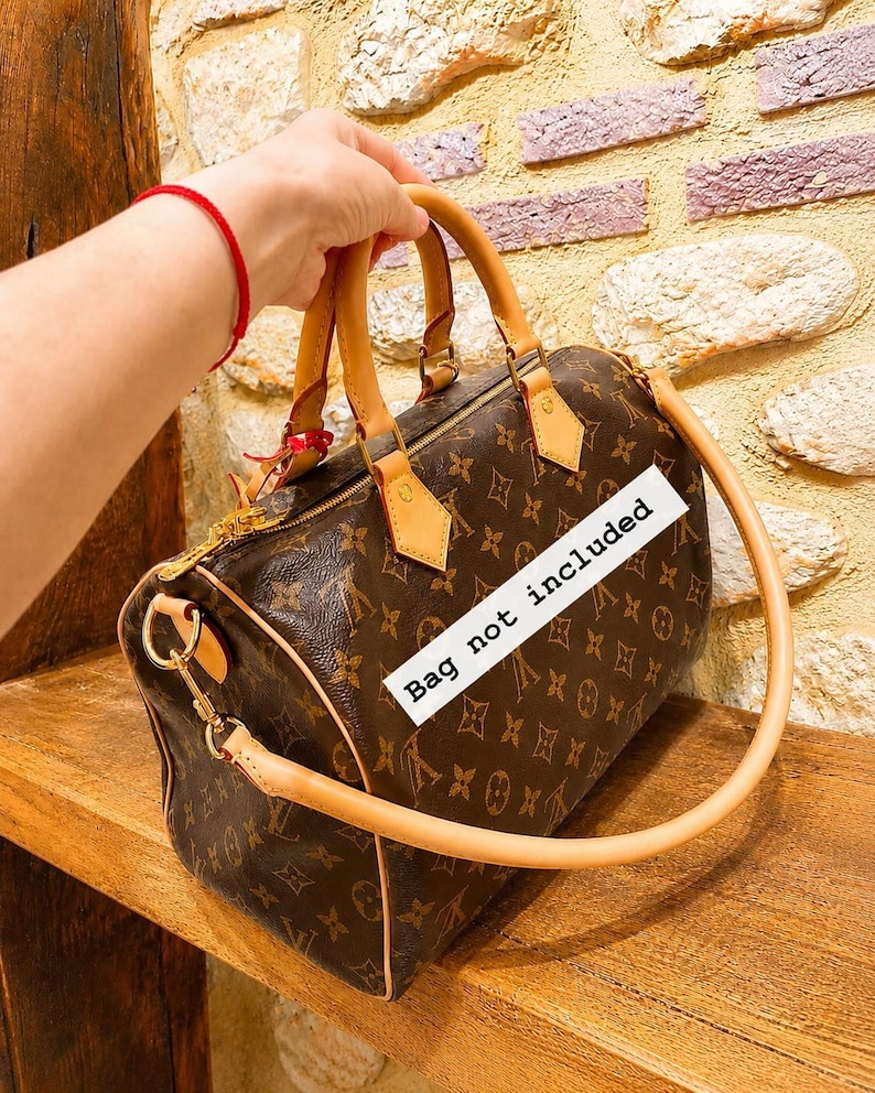 Pu&ograve; includere: Una borsa a mano marrone con un motivo monogramma marrone scuro ripetuto, manici in pelle color cuoio chiaro e dettagli in metallo dorato. Un'etichetta rettangolare bianca con il testo nero "Bag not included" &egrave; posizionata sulla borsa.
