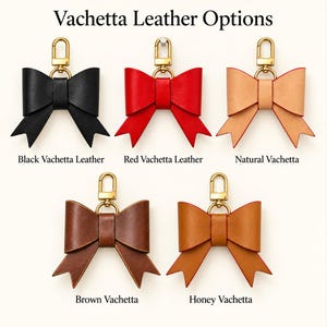 Könnte beinhalten: Fünf schleifenförmige Schlüsselanhänger aus Leder mit goldfarbenen Karabinerverschlüssen, beschriftet mit Vachetta Leather Options. Die Farben sind Schwarz, Rot, Natur, Braun und Honig. Jede Schleife hat ein Mittelband und zwei spitze Enden.