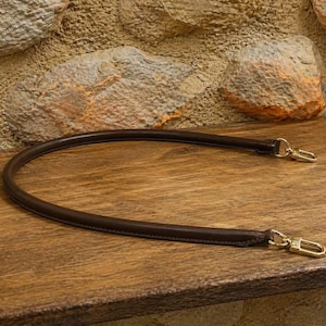 Pu&ograve; includere: Una tracolla per borsa in pelle marrone scuro, di forma arrotondata, dotata di chiusure a moschettone in metallo color oro lucido su entrambe le estremit&agrave;. Il cinturino &egrave; curvato a semicerchio, con cuciture lisce sui bordi e una struttura tubolare.