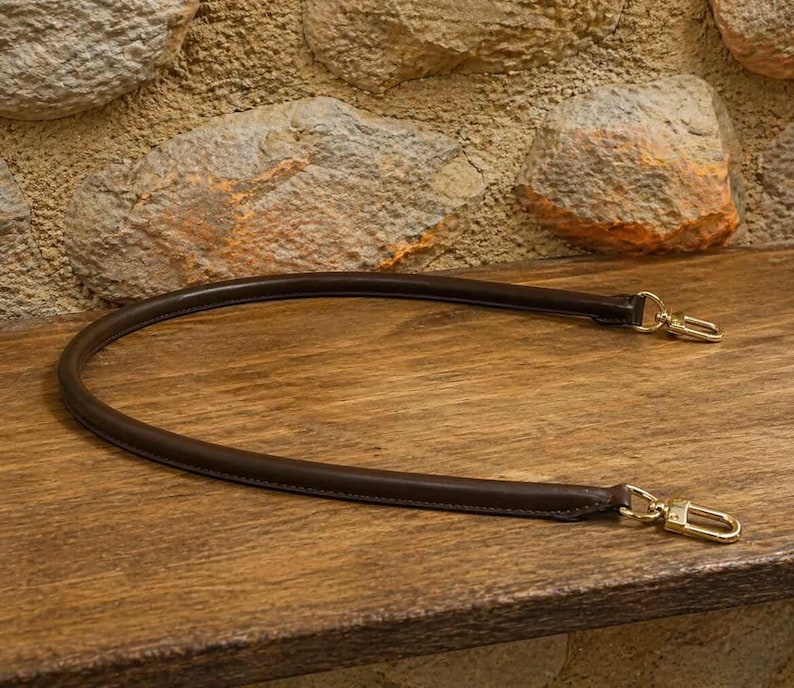 Pu&ograve; includere: Una tracolla per borsa in pelle marrone scuro, di forma arrotondata, dotata di chiusure a moschettone in metallo color oro lucido su entrambe le estremit&agrave;. Il cinturino &egrave; curvato a semicerchio, con cuciture lisce sui bordi e una struttura tubolare.