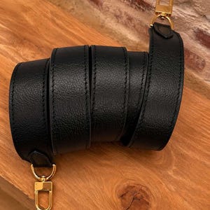 May include: A black leather strap with gold-colored metal clasps. The strap is rolled up, showcasing its texture and stitching. The clasps are attached at each end, ready for use. The background is a wooden surface.