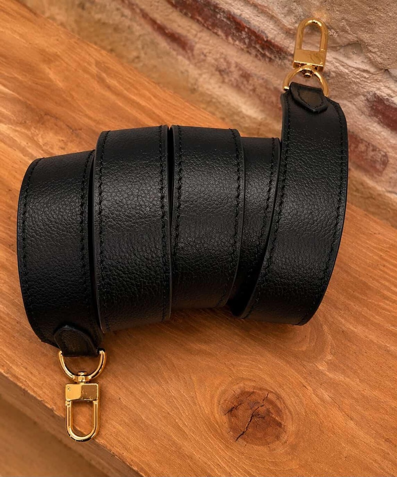 May include: A black leather strap with gold-colored metal clasps. The strap is rolled up, showcasing its texture and stitching. The clasps are attached at each end, ready for use. The background is a wooden surface.