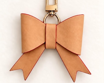 May include: A tan leather bow keychain with red edges and a gold-toned metal lobster clasp. The bow features a central leather band and two pointed tails. The text Vachetta Color is printed in brown below the bow on a plain white background.