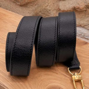 May include: A black leather strap with a gold-colored clasp. The strap is coiled on a wooden surface, showcasing its texture and stitching. The clasp is a shiny gold color.