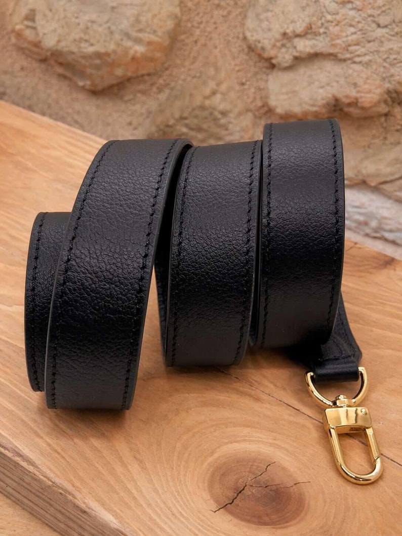May include: A black leather strap with a gold-colored clasp. The strap is coiled on a wooden surface, showcasing its texture and stitching. The clasp is a shiny gold color.