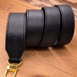 May include: A black leather strap with a gold-colored clasp. The strap is rolled up, showcasing its texture and stitching. The clasp is a shiny gold color, and the strap appears to be made of high-quality leather.
