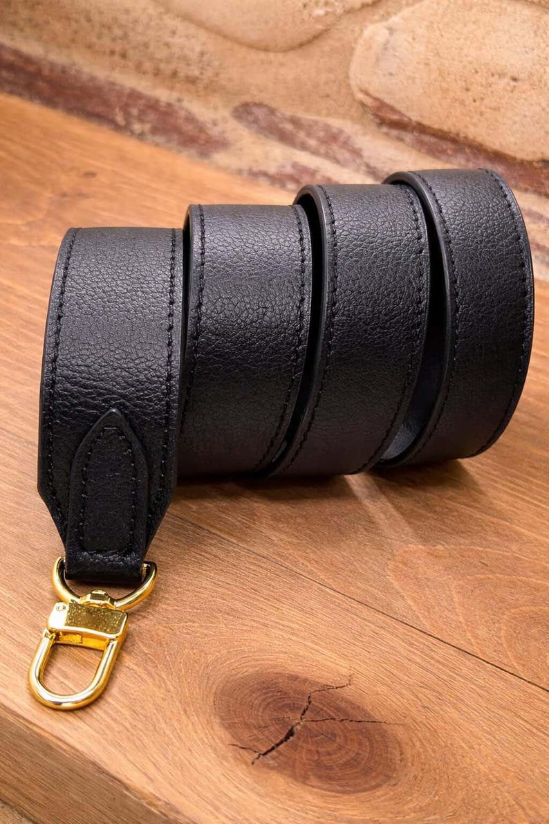 May include: A black leather strap with a gold-colored clasp. The strap is rolled up, showcasing its texture and stitching. The clasp is a shiny gold color, and the strap appears to be made of high-quality leather.