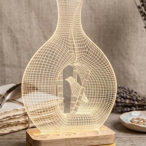 May include: An illuminated LED lamp with a bird and bamboo design inside a vase shape. The lamp has a grid pattern and rests on a wooden base with a yellow trim. The text "Deckrate01" is visible.