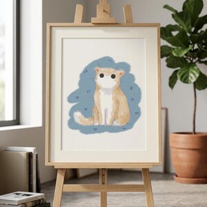 以下が含まれることがあります： 木製のイーゼルに飾られた、額入りの猫のアートプリント。アートワークは、青い背景に明るいオレンジと白の猫を描いています。イーゼルは明るい色の木でできており、明るい部屋に飾られています。