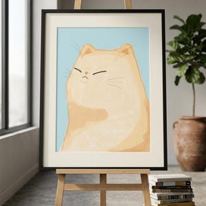以下が含まれることがあります： イーゼルに飾られた猫の額入りアートプリント。このアートワークは、水色の背景に目を閉じた明るいオレンジ色の猫を描いています。フレームは黒で、イーゼルは木製です。本が積み重ねられています。