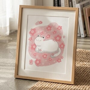 以下が含まれることがあります： ピンクの花柄の背景に、白い猫のイラストが描かれた額入りのアートプリント。猫はピンクの花に囲まれています。フレームは明るい色の木でできており、アートワークはニュートラルカラーのマットに表示されています。