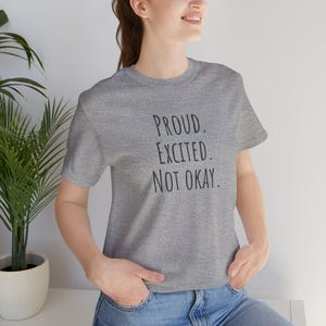 May include: A heather gray short-sleeve t-shirt featuring the black text "PROUD. EXCITED. NOT OKAY." printed in a simple, sans-serif font on the front. The garment has a crew neckline and a standard fit, constructed from a soft, heathered fabric.
