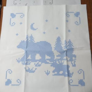 May include: White fabric with a blue cross-stitch design featuring a bear, trees, and a crescent moon. The design is part of a quilt block set, as indicated by the text "Black Bear Quilt Blocks - Set of 6" and the "Herrschners" logo.