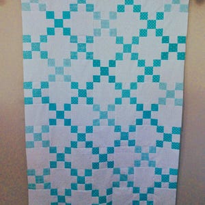 May include: A rectangular quilt featuring a repeating grid pattern of white squares and teal squares. The teal squares display various geometric patterns, including dots and cross-hatch designs, creating a consistent visual rhythm across the fabric.