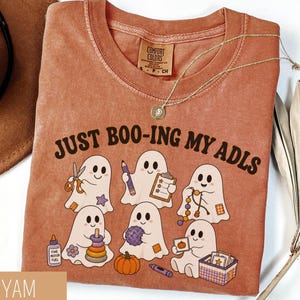 May include: A rust-colored t-shirt featuring a graphic of six white ghosts engaged in various activities. The text "JUST BOO-ING MY ADLS" is printed above the ghosts. The shirt has a crew neckline and a Comfort Colors brand label at the neck.