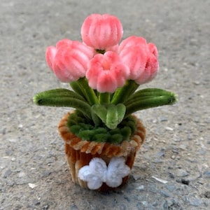 May include: A miniature handcrafted flower arrangement featuring pink tulips and green leaves in a brown pot with a white butterfly accent. The flowers are made of a soft, fuzzy material, creating a charming, whimsical aesthetic. The pot is approximately 3 inches tall.