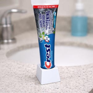May include: A blue and white tube of Crest toothpaste with the text "Baking Soda & Peroxide Whitening" and "Fresh Mint". The toothpaste is in a white holder on a bathroom countertop.